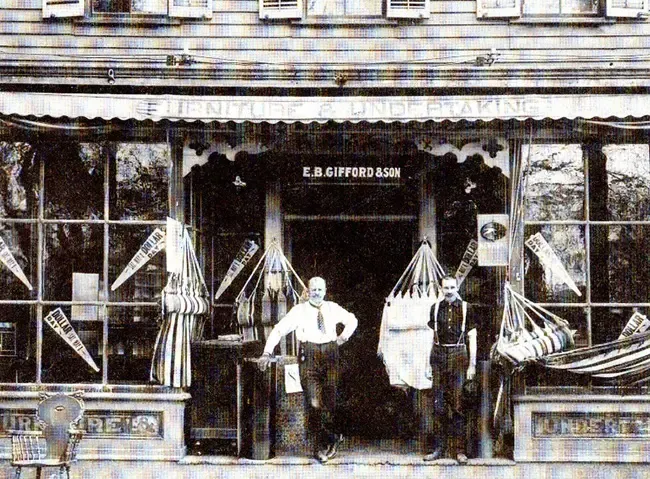 Storefront with two men; awnings, signs, and fabric displays; old-fashioned building.