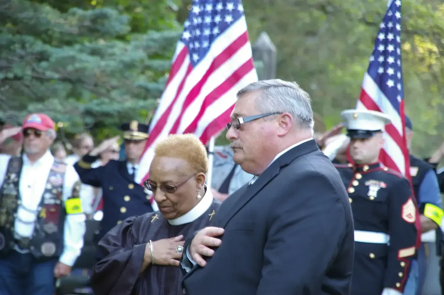 People at a memorial with US flags. Man in suit, woman in religious attire, others salute.