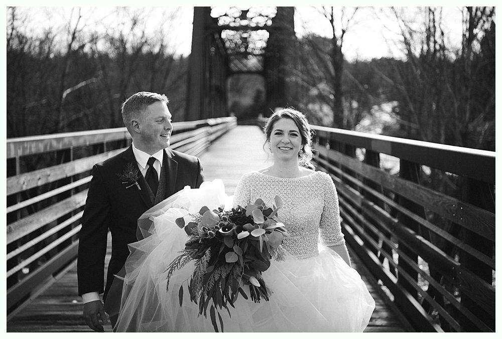 Bride and groom walk hand-in-hand across a wooden bridge, holding a bouquet. Black and white.
