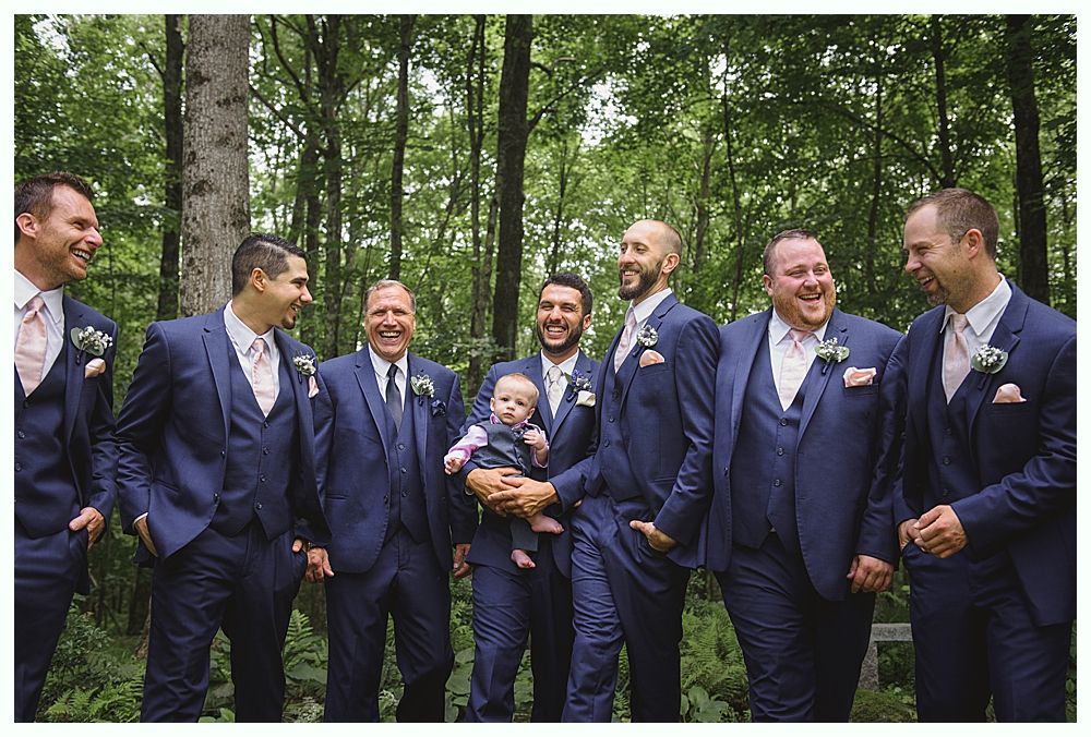 Groomsmen in dark suits pose on a grassy hillside under a cloudy sky.