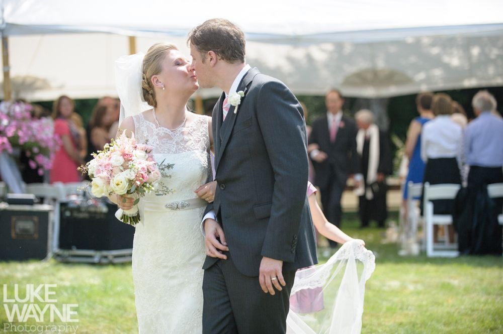 Newlyweds kiss, arm in arm, exiting wedding ceremony under a tent. Bride holds bouquet, groom in suit.