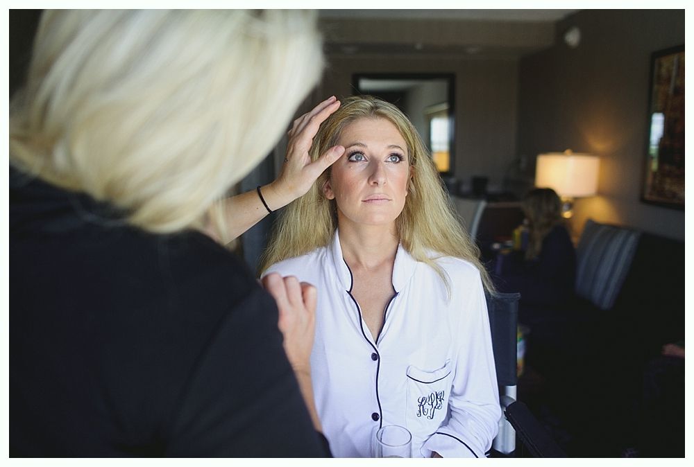Woman having makeup applied in a hotel room, wearing a white shirt; makeup artist's hands near her face.