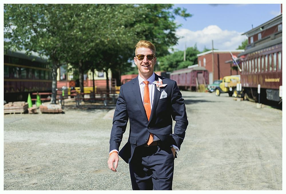 Man in navy suit and orange tie walks toward the camera; background of train cars and buildings.