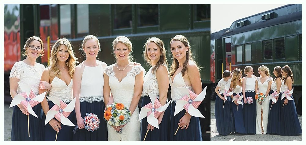 Bridesmaids in navy skirts and white tops hold pinwheels by a train. The bride holds a bouquet.