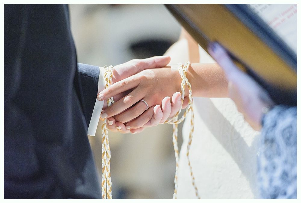 Wedding ceremony: hands clasped, tied with gold cords, ring visible. Ceremony officiant holding book.