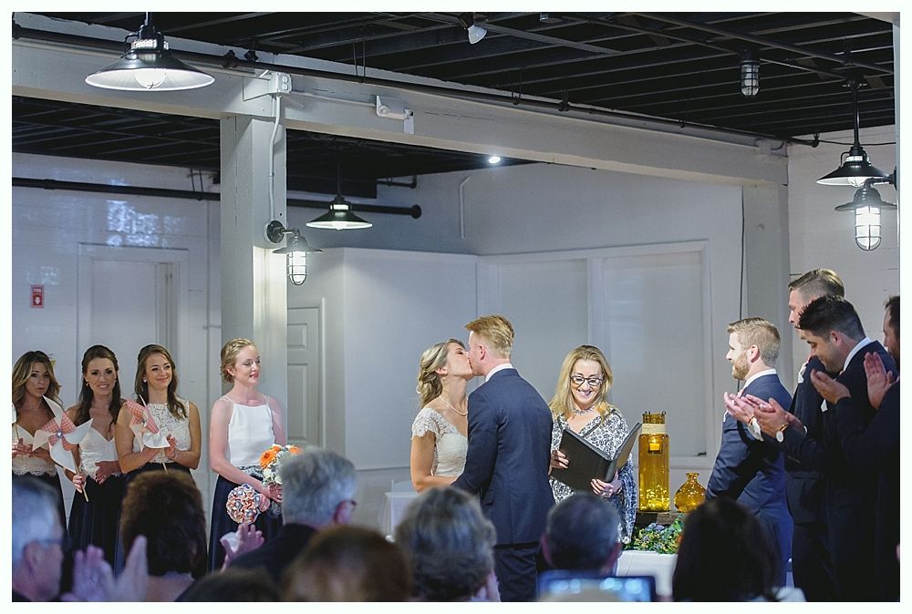 Couple kissing at wedding ceremony, surrounded by wedding party and guests. Indoor setting with industrial-style lighting.