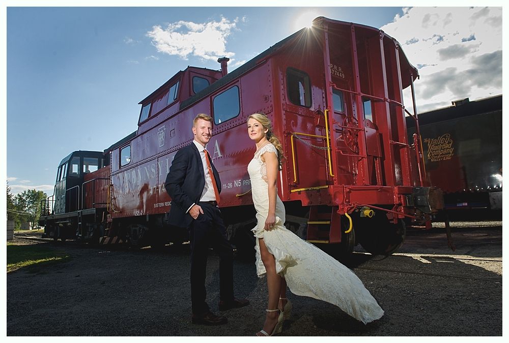 Couple poses by a red caboose on a sunny day. The bride wears a white dress; the groom, a dark suit.