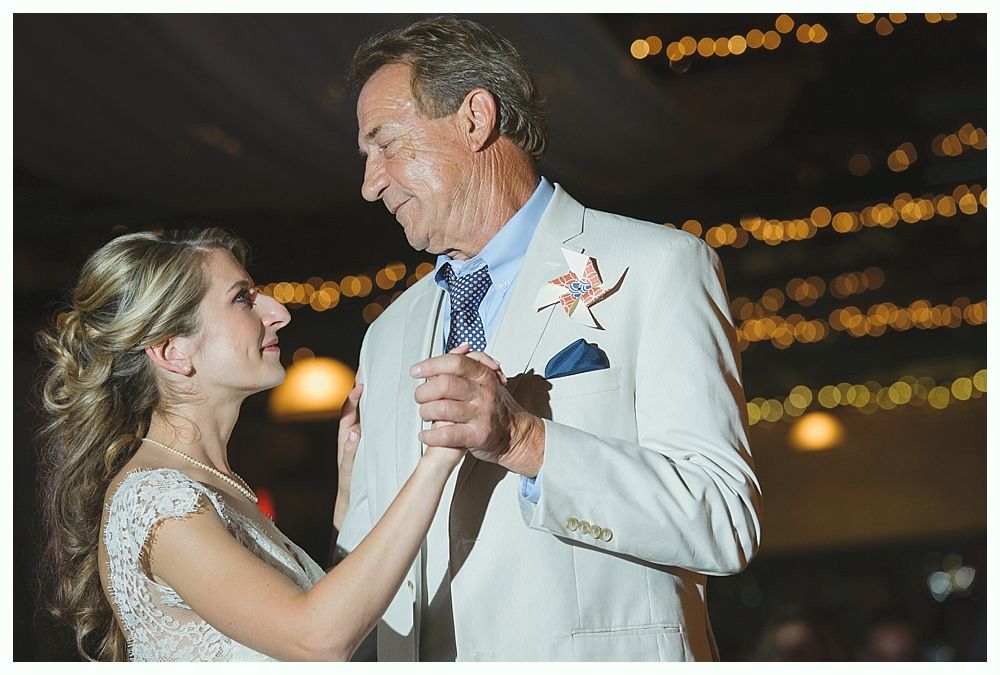 Bride and father dance at wedding reception. They look at each other, smiling. Gold lights are in the background.