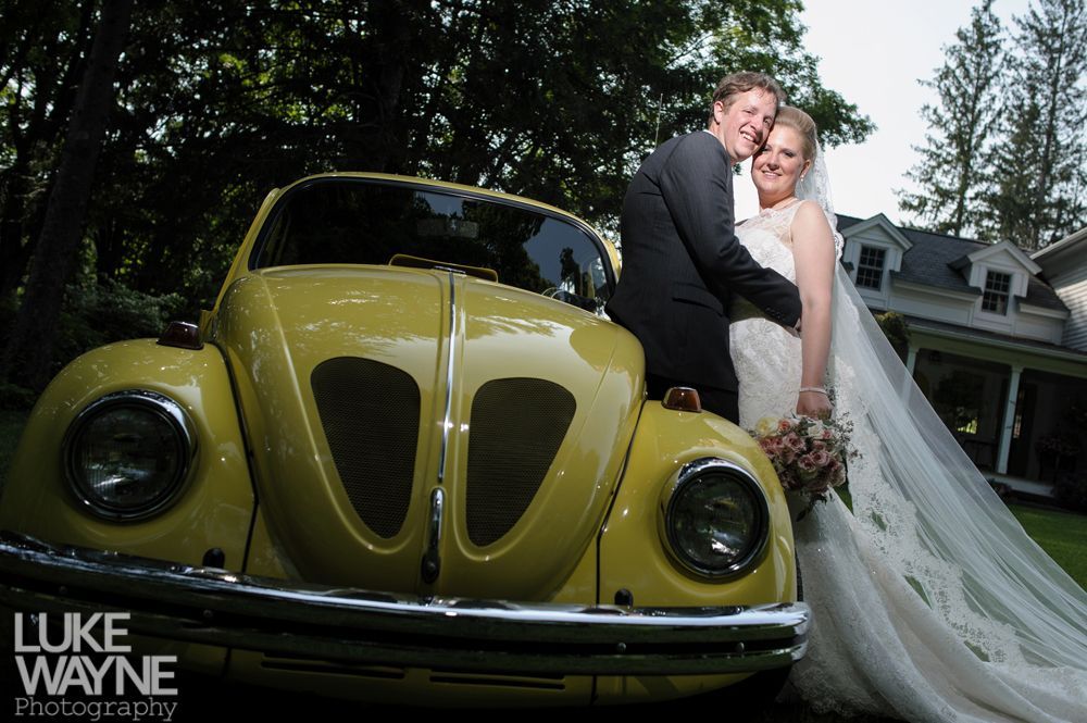 Bride and groom embrace by a yellow VW Beetle. She wears a white gown, veil, and holds flowers.
