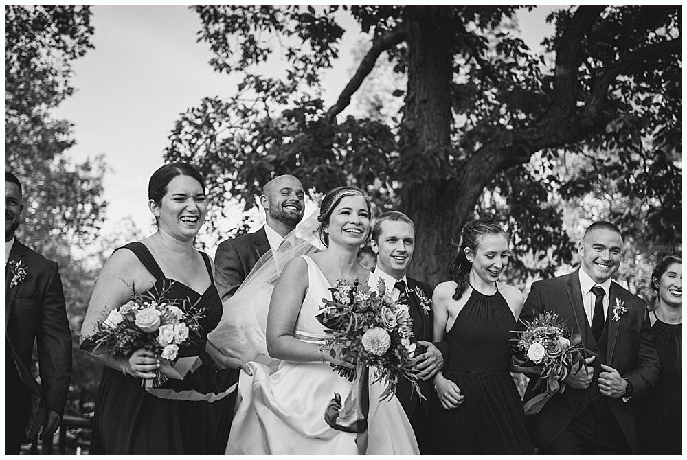 Newlyweds exiting outdoor wedding ceremony, holding hands, smiling. Wedding party applauds.