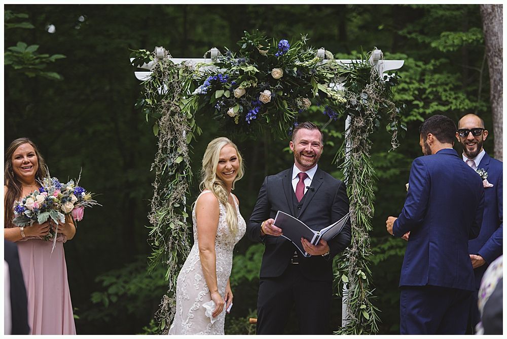 Wedding party poses outdoors. Groomsmen in navy suits, bridesmaids in red dresses, bride in white dress, holding bouquets.