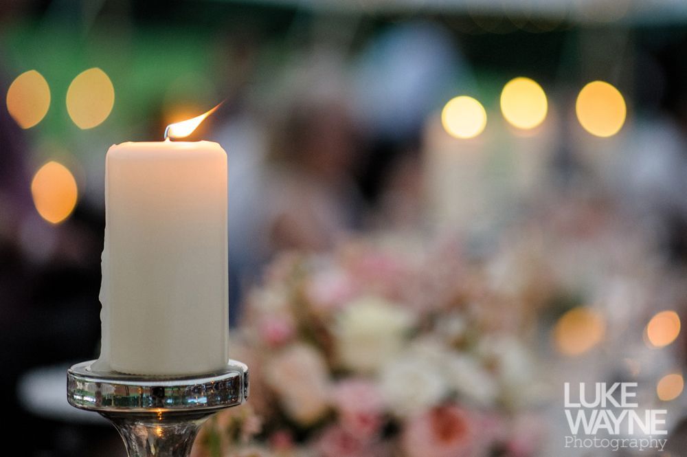 Lit white candle in silver holder, blurry background of flowers and soft lights.
