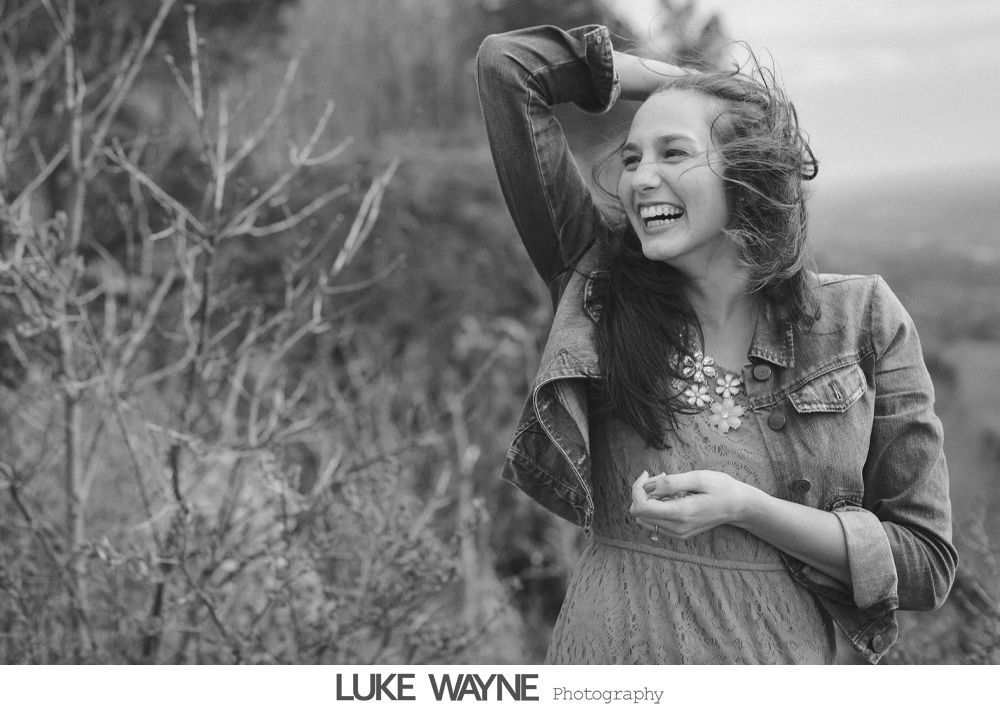 Woman laughs joyfully outdoors, hand in hair, wearing denim jacket and floral dress.