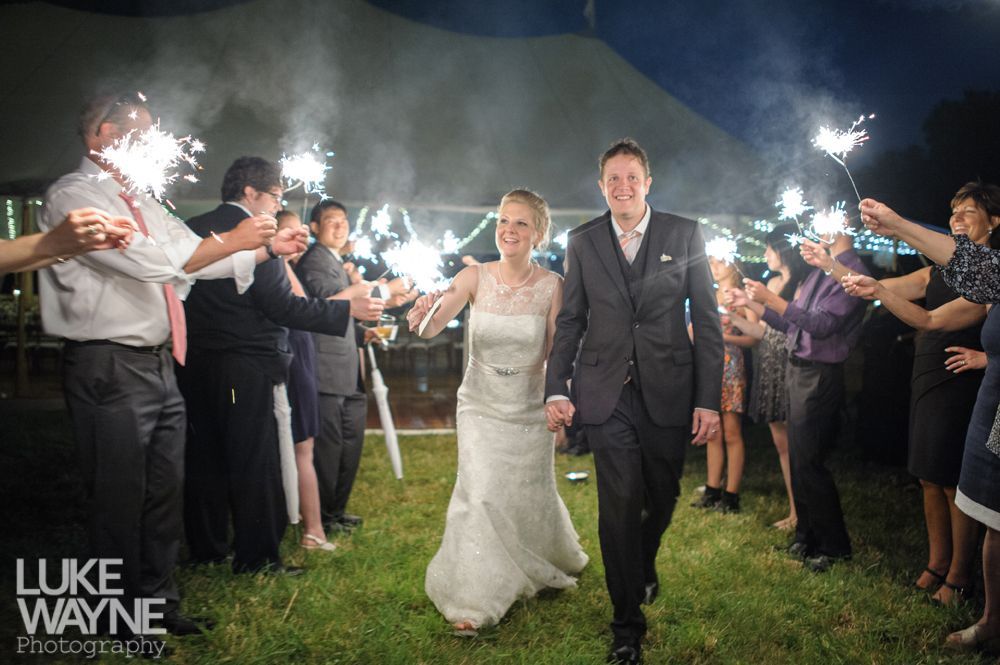 Bride and groom walk through sparkler-lined path, smiling. Wedding guests cheer under a tent at dusk.