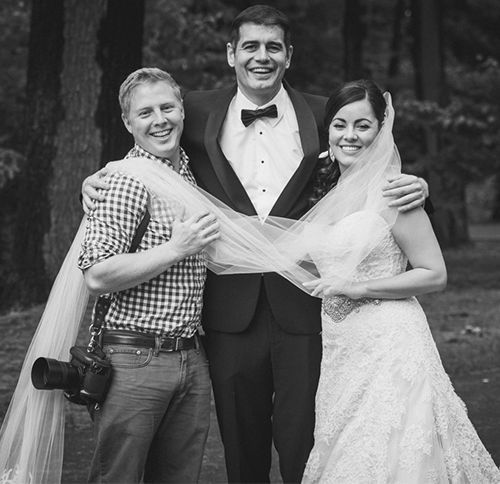 A wedding photographer poses with the smiling bride and groom, holding her veil. Black and white.