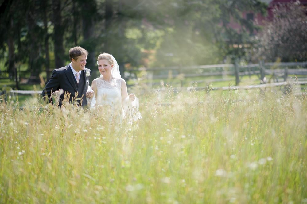 Bride and groom walking through a tall grassy field, smiling. Wooden fence and trees in background.
