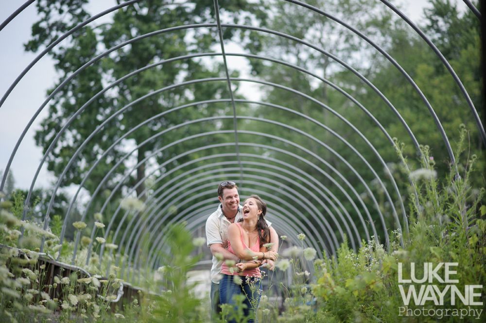 Couple embraces beneath arched metal structure in a garden, smiling, with flowers and greenery.