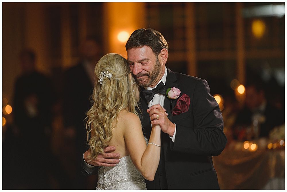 Bride and groom embrace outdoors, veil billowing, wedding bouquets visible.