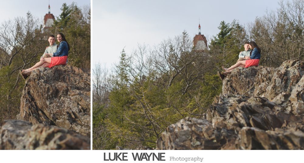 Couple sits on a rocky outcrop, distant building visible. He wears a blue jacket, she wears an orange skirt. Outdoors.