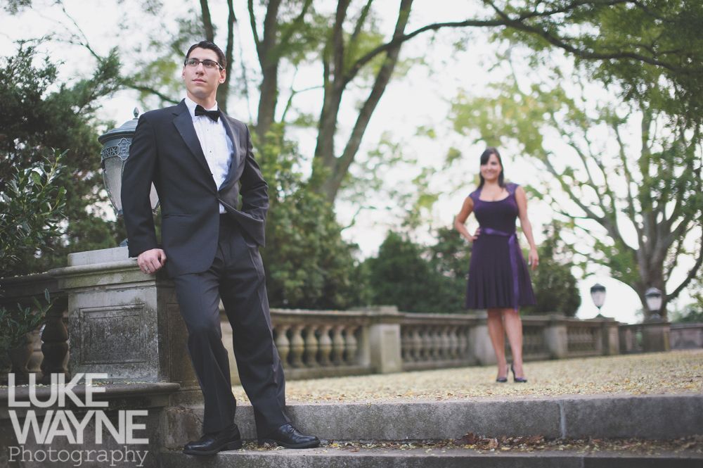 Man in tuxedo poses on steps, woman in purple dress stands behind him on a terrace.