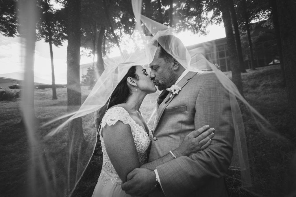 Bride and groom kissing under a veil outdoors. Sunlight filters through trees. Black and white.
