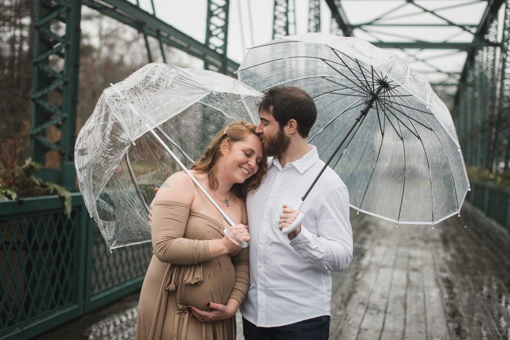 Pregnant couple under clear umbrellas on a bridge; man kisses woman's forehead, both smiling.