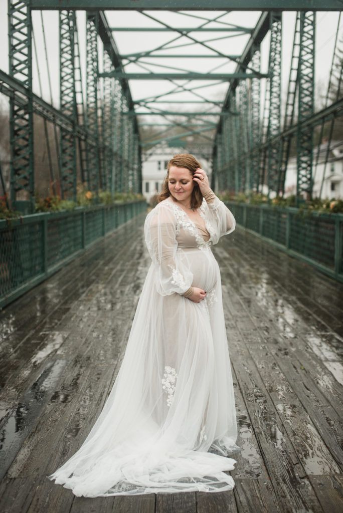 Pregnant person in white gown on a bridge, smiling, holding belly. Bridge is green, raining, outdoors.