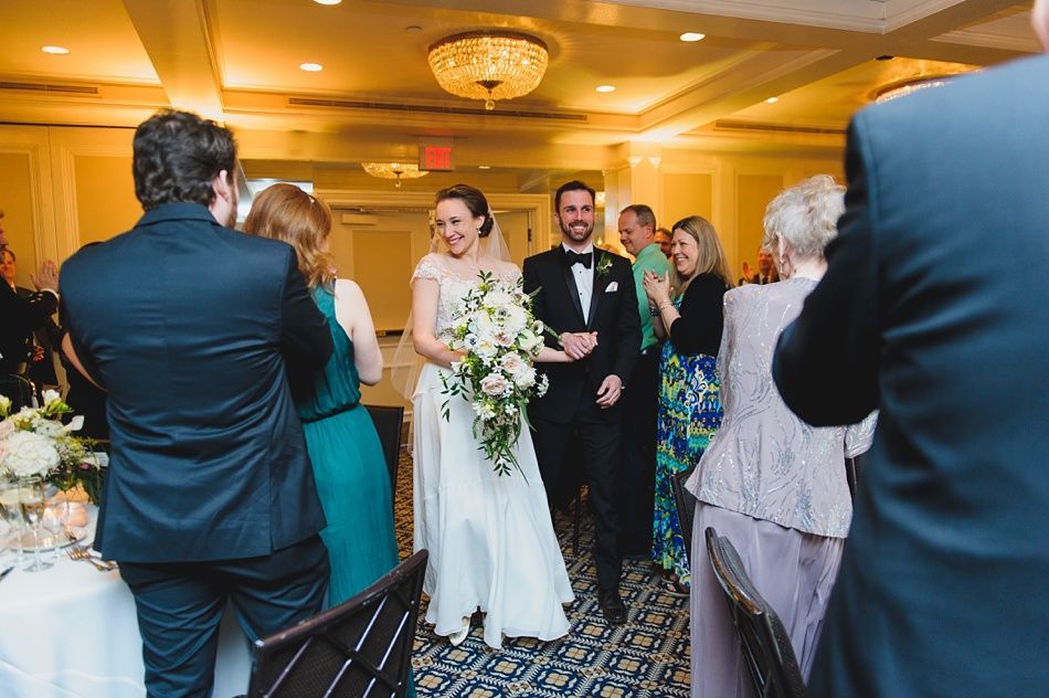 Bride with arms up, making a funny face, groom looking at his hand, cake cutting in background.
