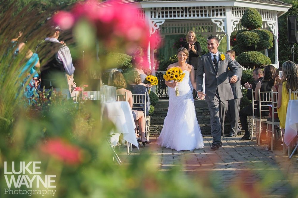 Newlyweds exiting outdoor ceremony, holding hands, with guests, gazebo, and flowers.