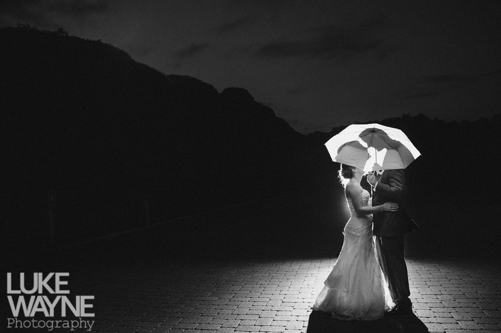 Wedding couple silhouetted under umbrella, lit from behind, against a dark mountain backdrop.