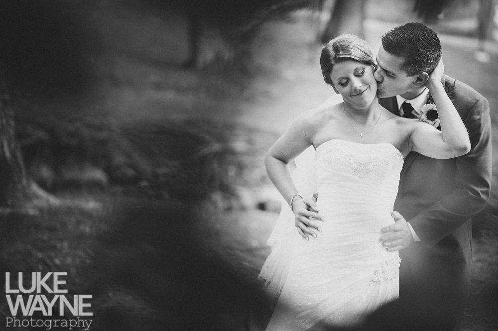 Bride and groom embrace outdoors, groom kissing bride's neck; black and white photo.