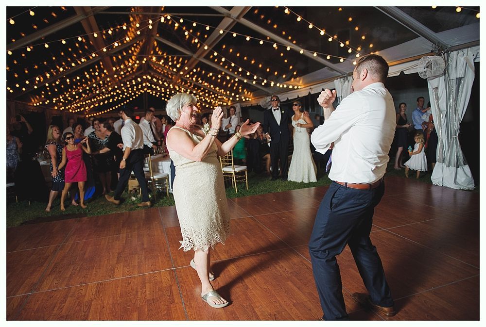 Bride with arms up, making a funny face, groom looking at his hand, cake cutting in background.