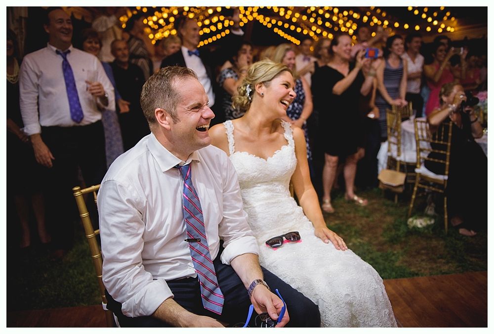 Bride with arms up, making a funny face, groom looking at his hand, cake cutting in background.