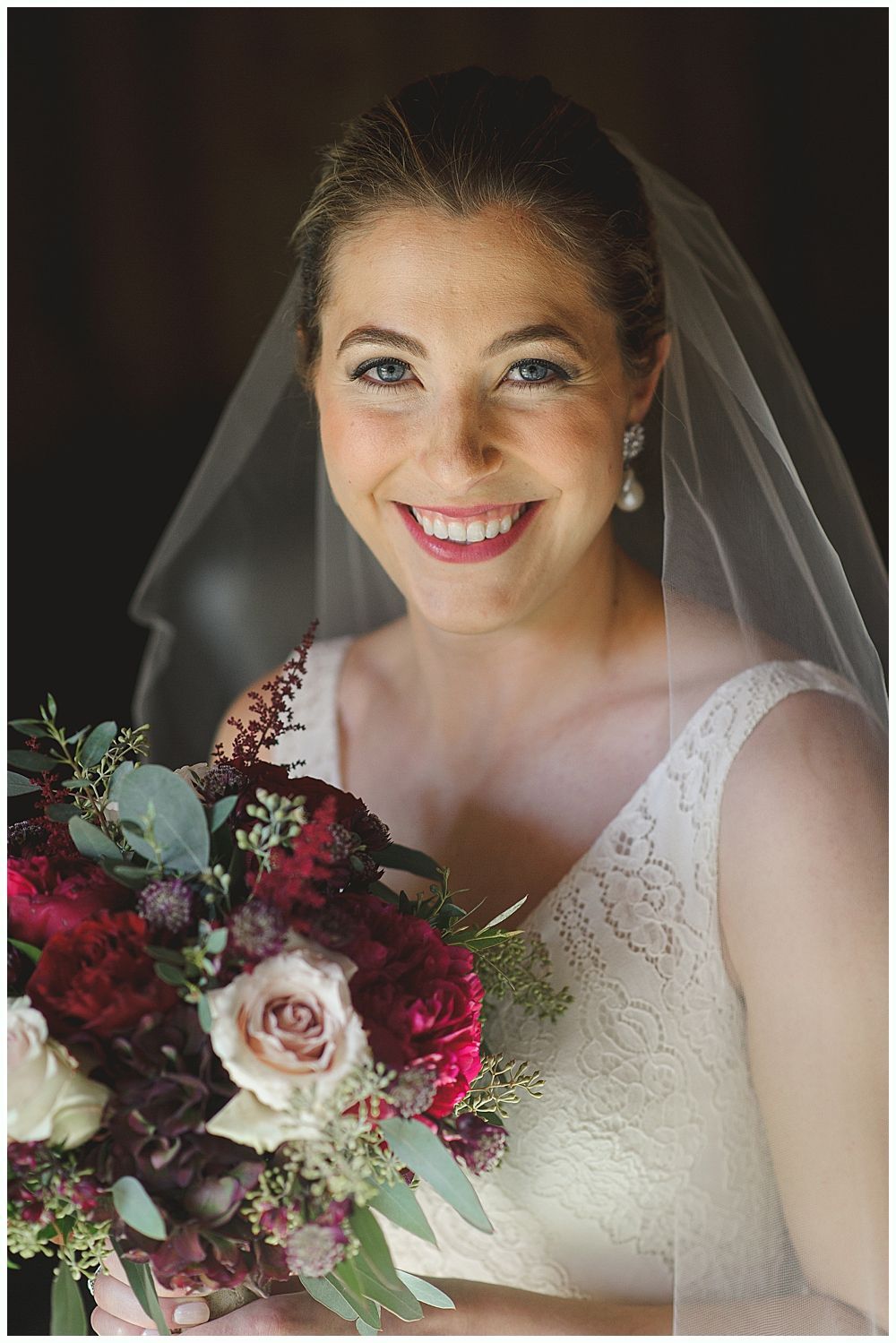Bride in a white lace dress with veil, smiling, holding a bouquet of burgundy and green flowers.