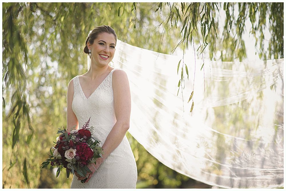Bride in a white wedding dress smiles, holding a bouquet. A veil flows in the wind under a weeping willow.