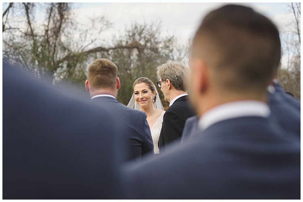 Bride smiles at the altar during a ceremony. Groomsmen in suits partially block view. Outdoor setting.