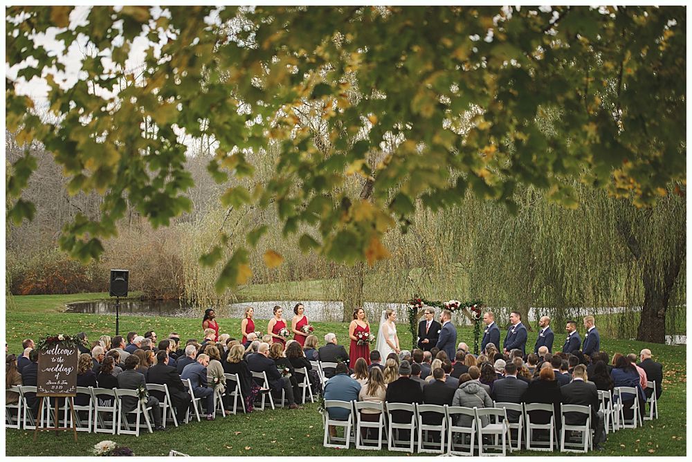 Wedding ceremony outdoors under a tree, guests seated. Couple, bridal party at altar. Autumn foliage.