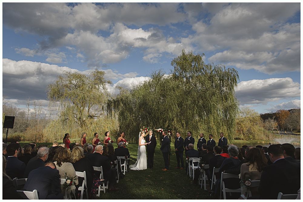 Wedding ceremony outdoors by a lake with a couple, wedding party, and guests under a cloudy sky.