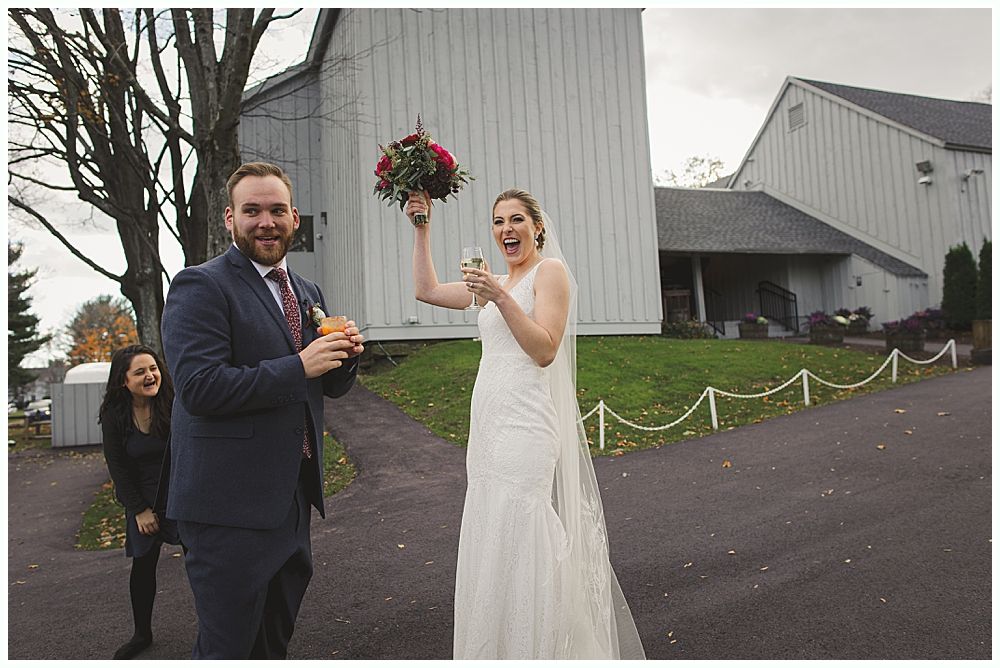 Newlyweds celebrating with champagne outside a building. Bride holding bouquet and bottle. Another person laughs in the background.