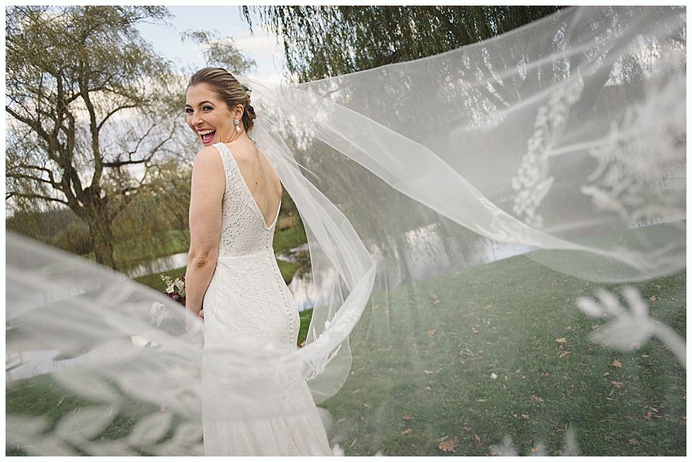 Bride in a white wedding dress smiles, looking over her shoulder as her veil blows in the wind outdoors.