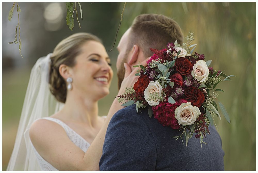 Bride touches groom's face; he holds a bouquet. They smile outdoors.