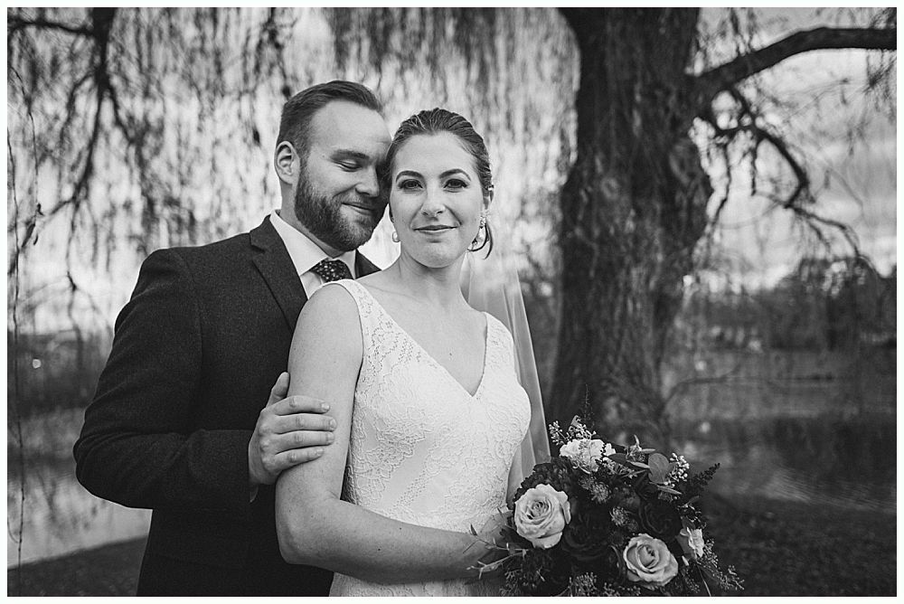 Bride and groom pose near a tree and pond. Bride in lace dress holds bouquet. Groom embraces her from behind.