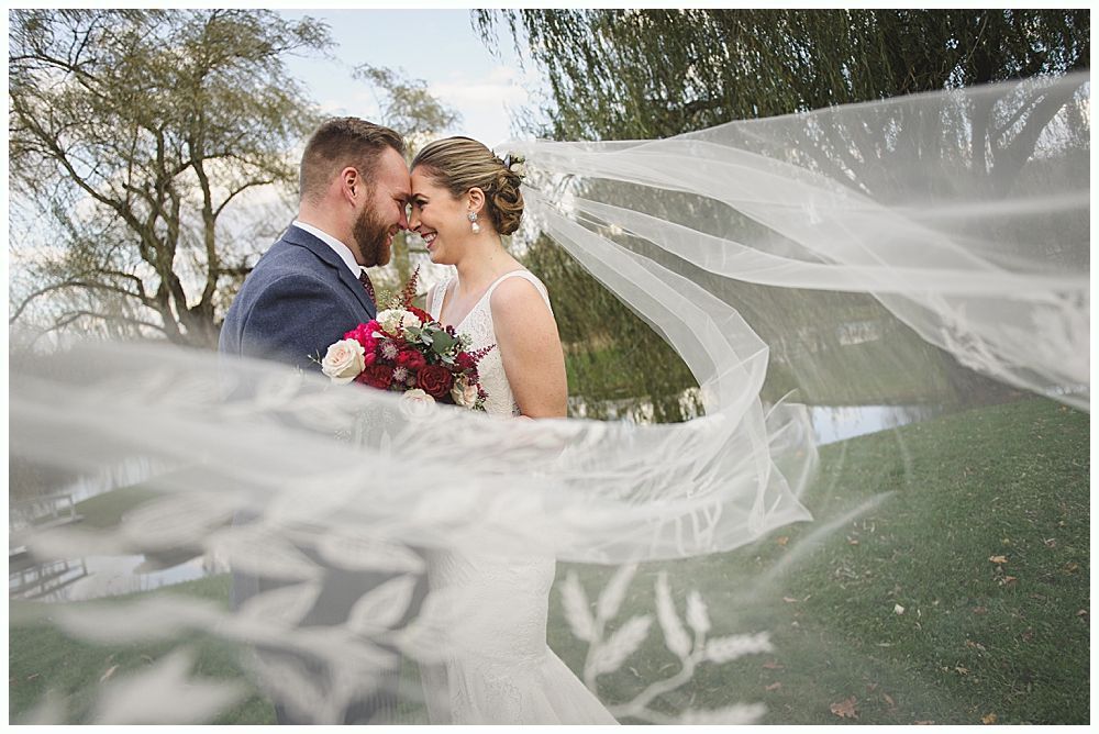 Bride and groom embrace outdoors, veil billowing, wedding bouquets visible.