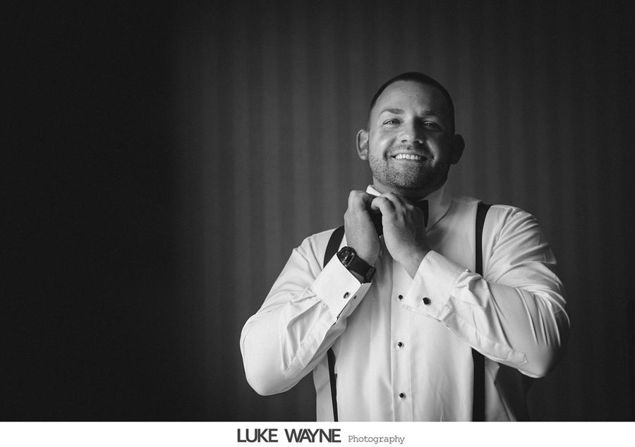 Groom adjusting bow tie, smiling. Wearing suspenders, dress shirt, in dim room with textured wall.