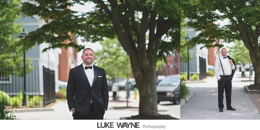 Two men in formal wear pose near a tree-lined street. One smiles in a black suit, other in suspenders, white shirt.