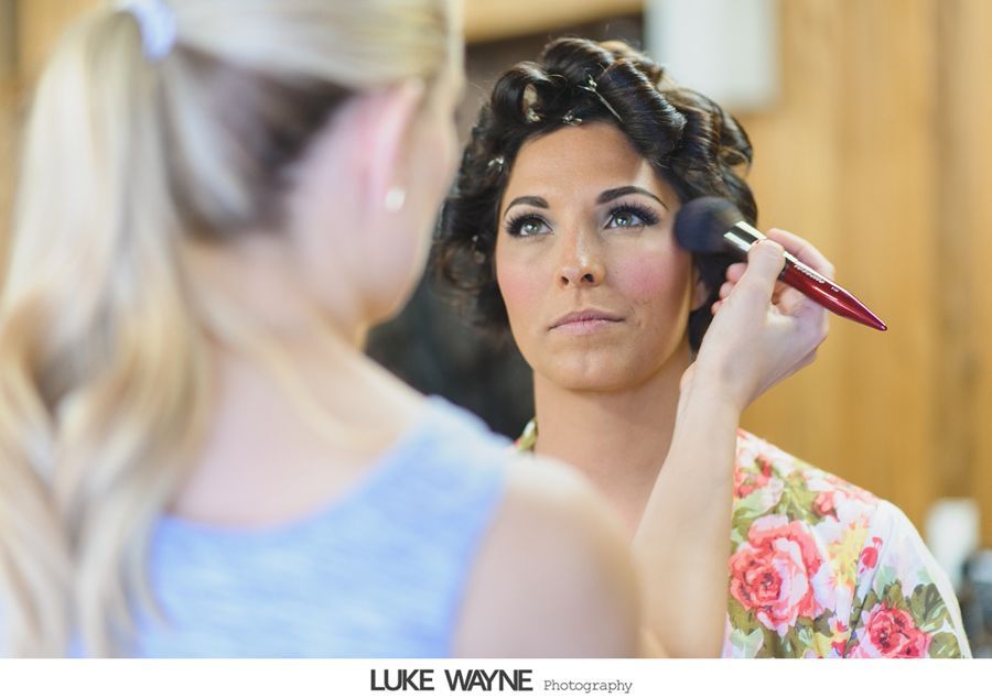 Makeup artist applying blush to a woman's face, wearing floral robe, hair in rollers, in a room with wood paneling.