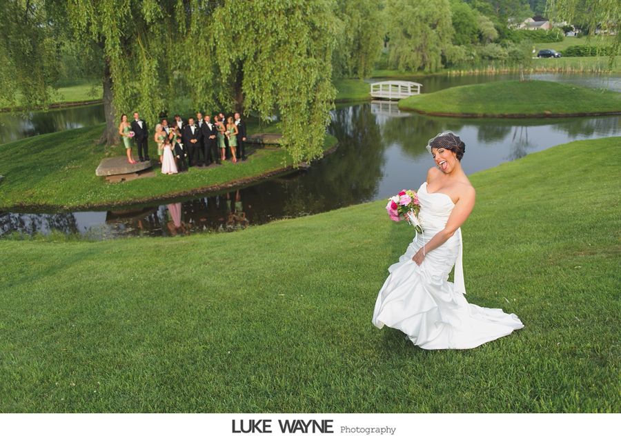 Bride in white gown poses on a grassy hill, wedding party by a pond under a tree.