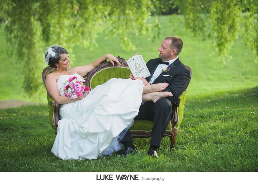 Bride and groom relax on green couch outdoors, bride's feet on groom's lap, holding card, smiling. Green backdrop.