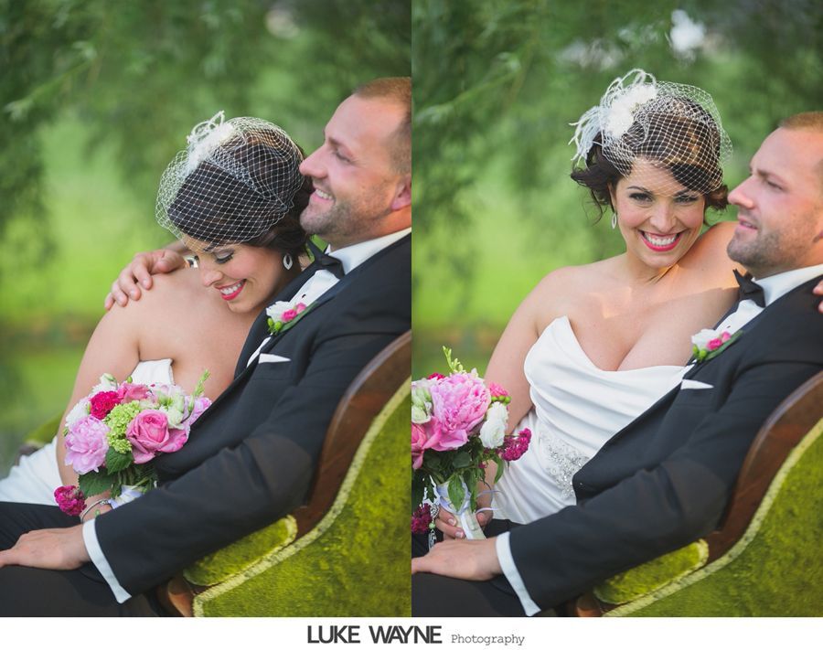 Bride and groom seated, bride in white strapless dress, groom in black suit, both smiling, sitting on a green bench outdoors.