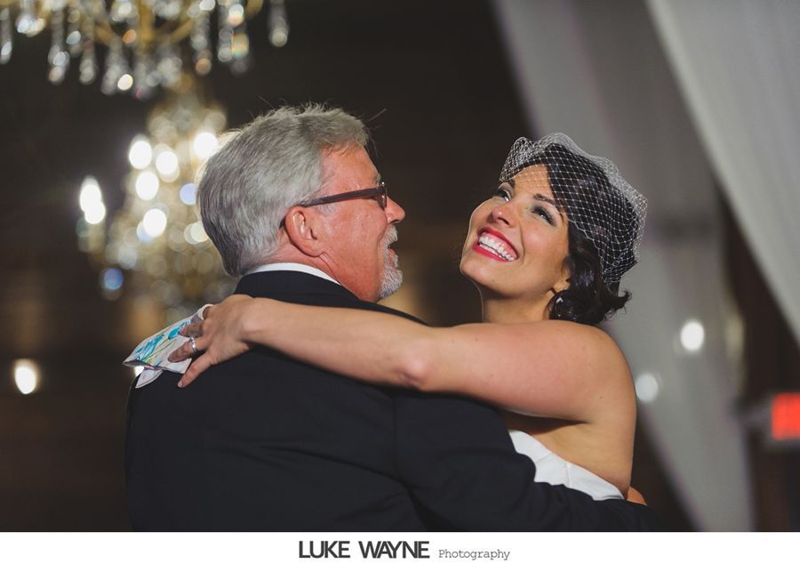 Bride and her father dance, bride smiling, indoors with chandelier.